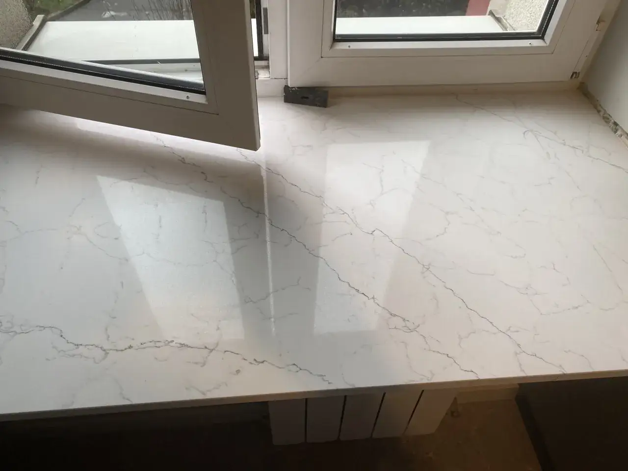 Biały parapet z konglomeratu z subtelnymi szarymi żyłkami, imitującymi marmur, pod otwartym oknem.