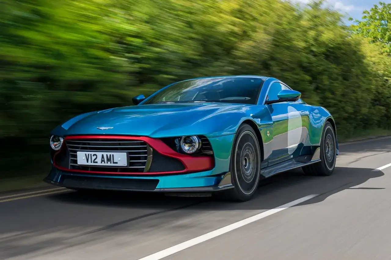 Ein blauer Aston Martin Valour rast auf einer Landstraße, umgeben von grünen Bäumen.