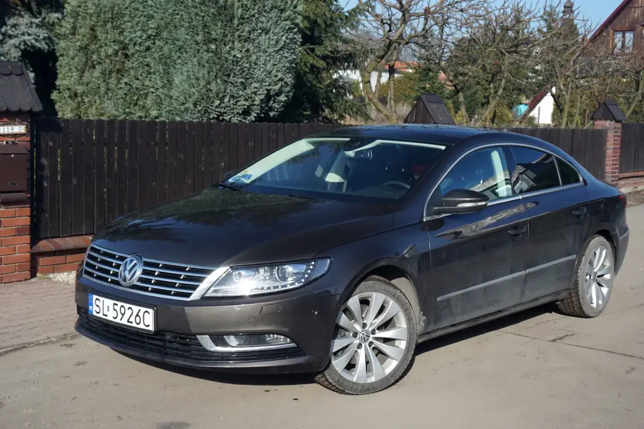 Ciemny Volkswagen Passat CC z tablicą SL 5926C zaparkowany przed drewnianym płotem i domem.