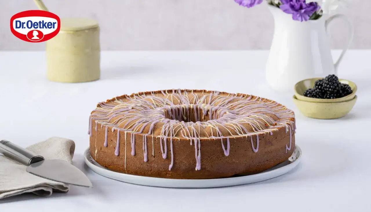 Ein **schneller Brombeerkuchen** mit lila und weißer Glasur, dekoriert mit frischen Brombeeren und Blumen.