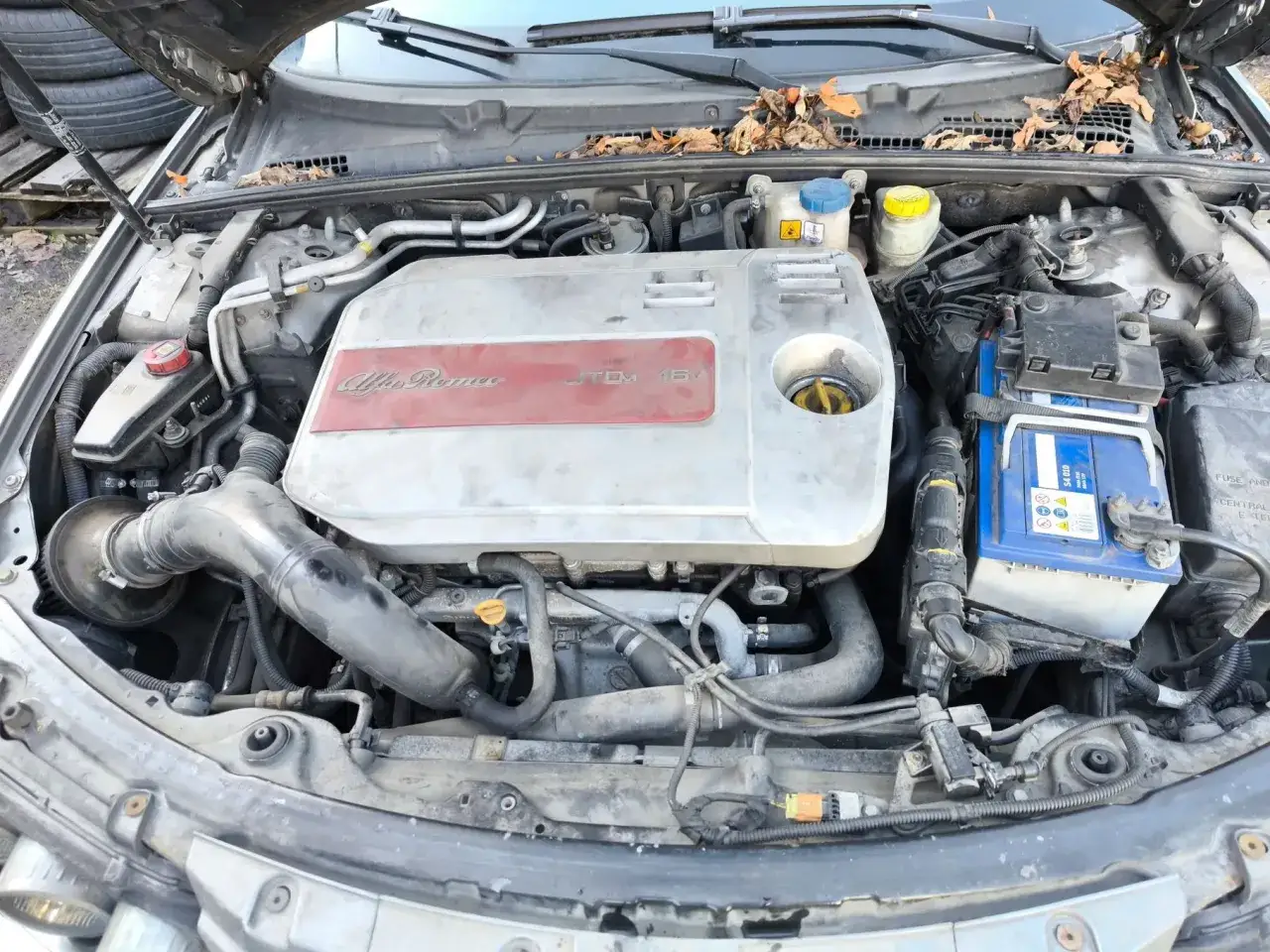 Silnik Alfa Romeo 159 1.9 JTD, z widocznym osprzętem i akumulatorem. Na pokrywie silnika widać napisy 
