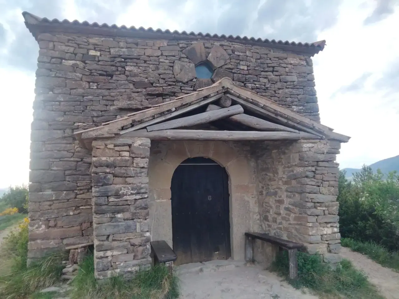 Las ermitas de Tella, construidas en piedra, invitan a la contemplación. Una puerta oscura y un banco de madera esperan bajo un tejado rústico.