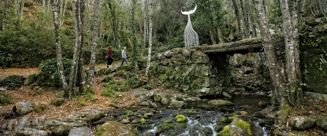 Dos personas pasean por el camino del agua en Cercedilla, junto a un arroyo y una escultura.