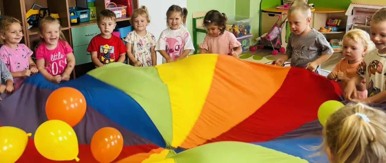 Wesołe dziecko w przedszkolu bawi się kolorowym spadochronem i balonami.