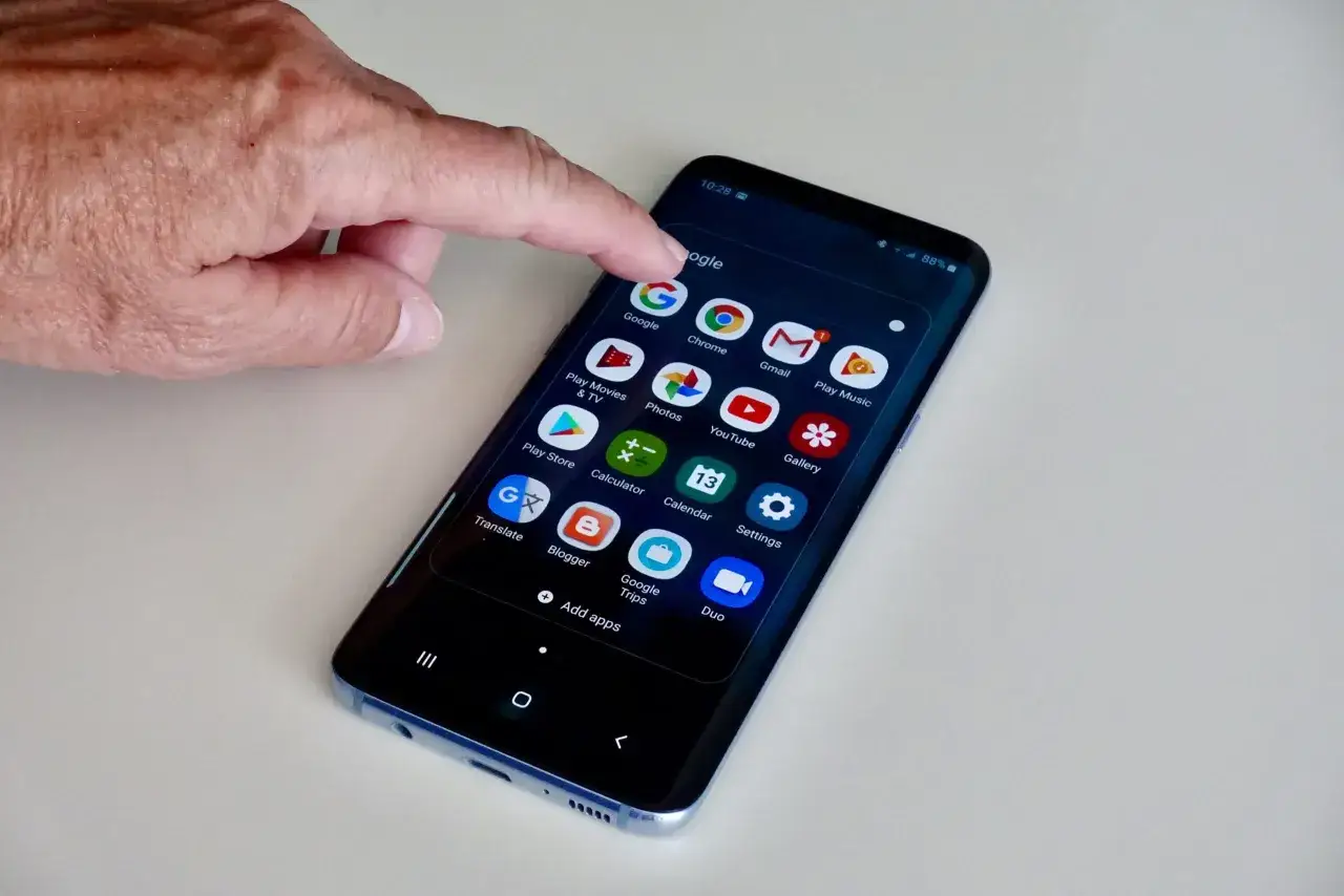 Dłoń naciska ikonę Google na ekranie smartfona z systemem android 13. Widoczne aplikacje: Chrome, Gmail, YouTube.