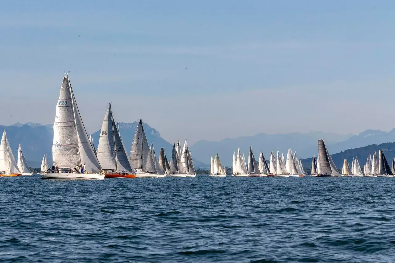 Viele Segelboote kämpfen bei einer Regatta um die Führung. Die Boote sind auf dem blauen Wasser, mit Bergen im Hintergrund.