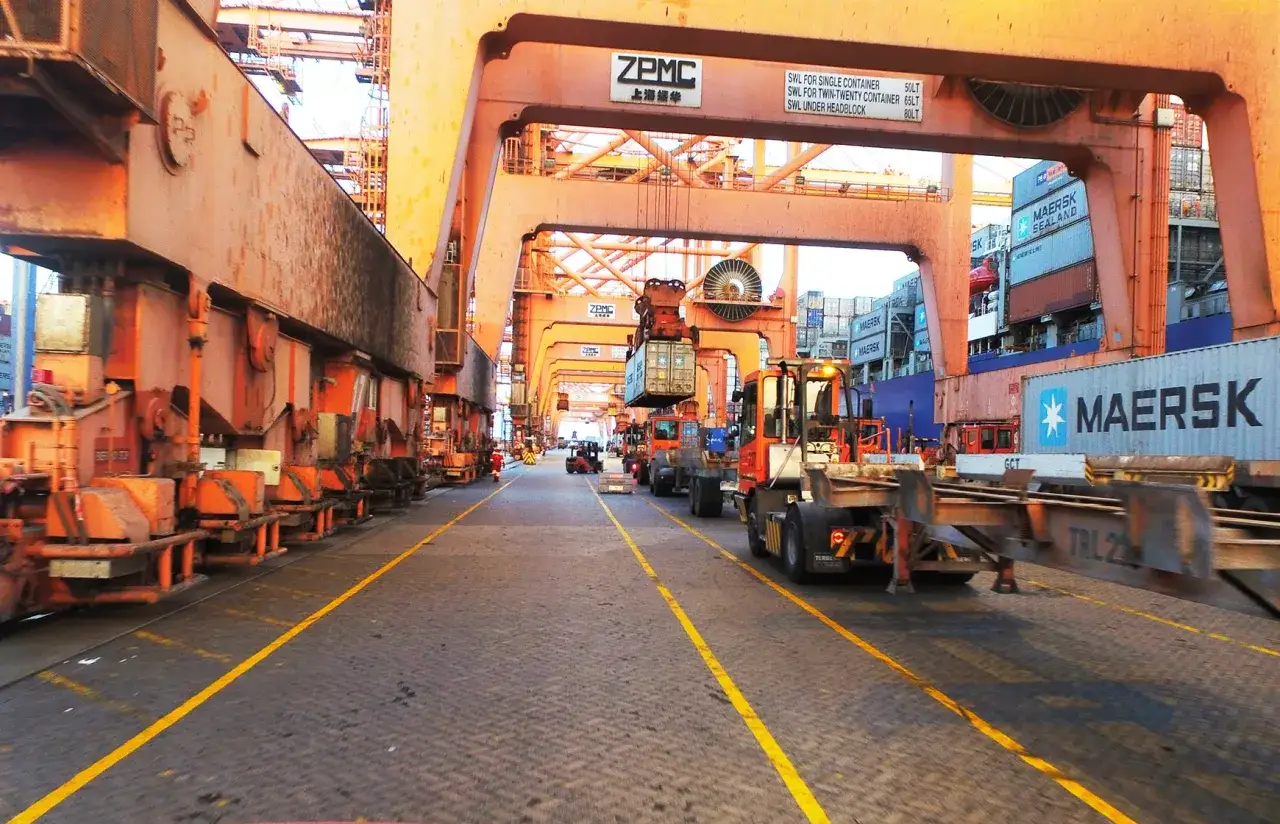 Große Kräne und Container im Hafen von Salalah. Ein Gabelstapler transportiert einen Container, während ein Schiff im Hintergrund beladen wird.