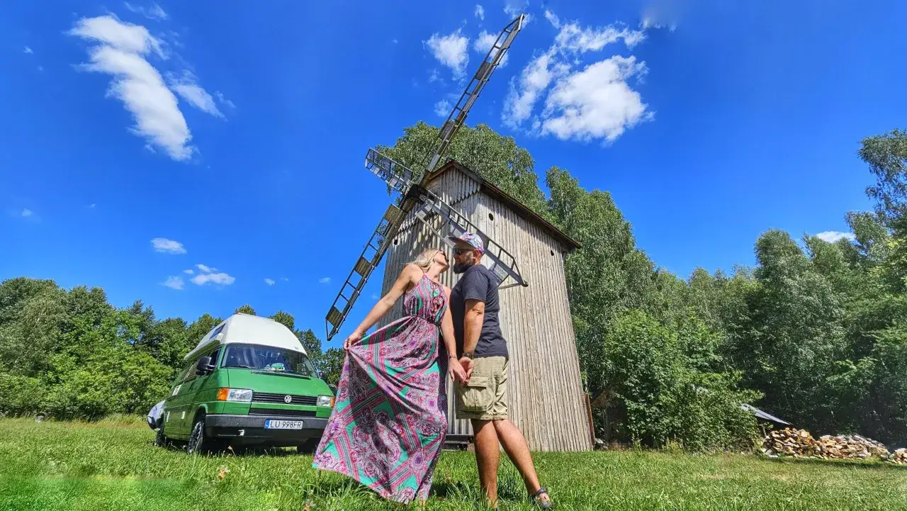 Para całuje się przy wiatraku na tle zieleni i błękitnego nieba. Obok stoi zielony kamper. Romantyczny wypad na Polesie Lubelskie.