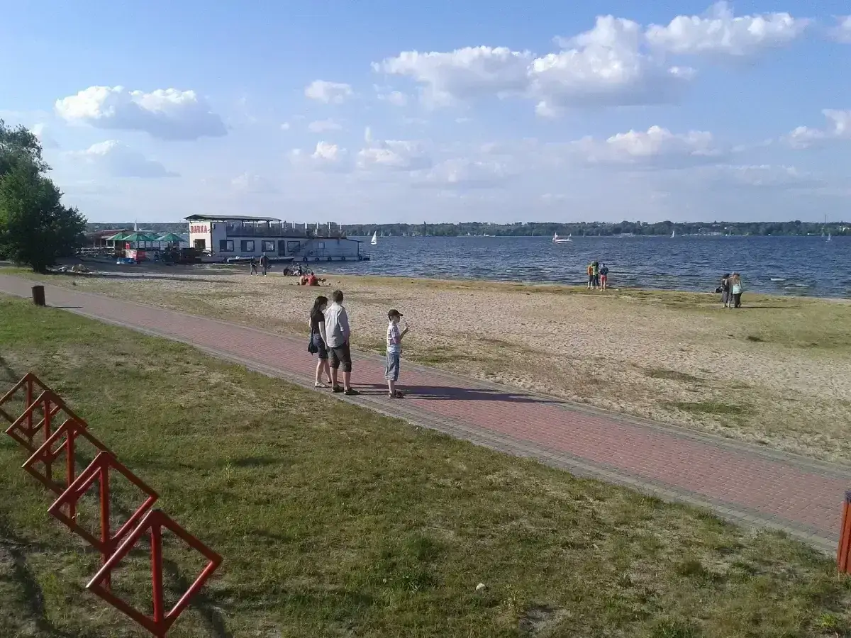 Rodzina spaceruje brzegiem jeziora Sierakowo, gdzie cumuje barka z restauracją. W tle żaglówki na wodzie.