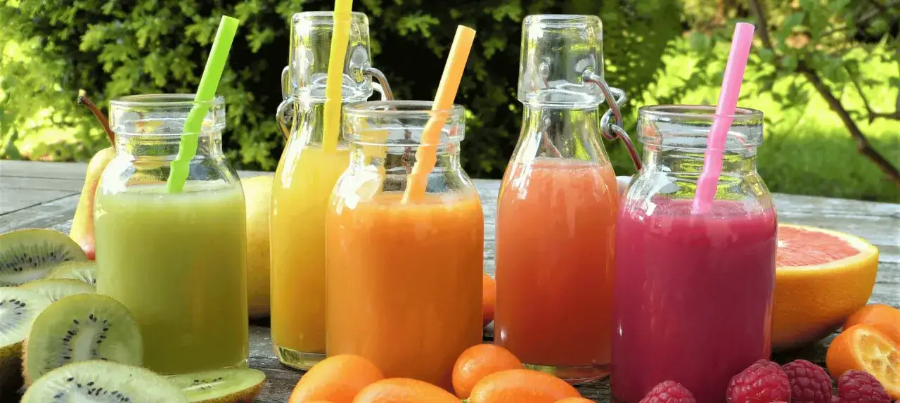 Kolorowe soki do drinków w butelkach, otoczone świeżymi owocami: kiwi, pomarańcze, maliny i grejpfrut.