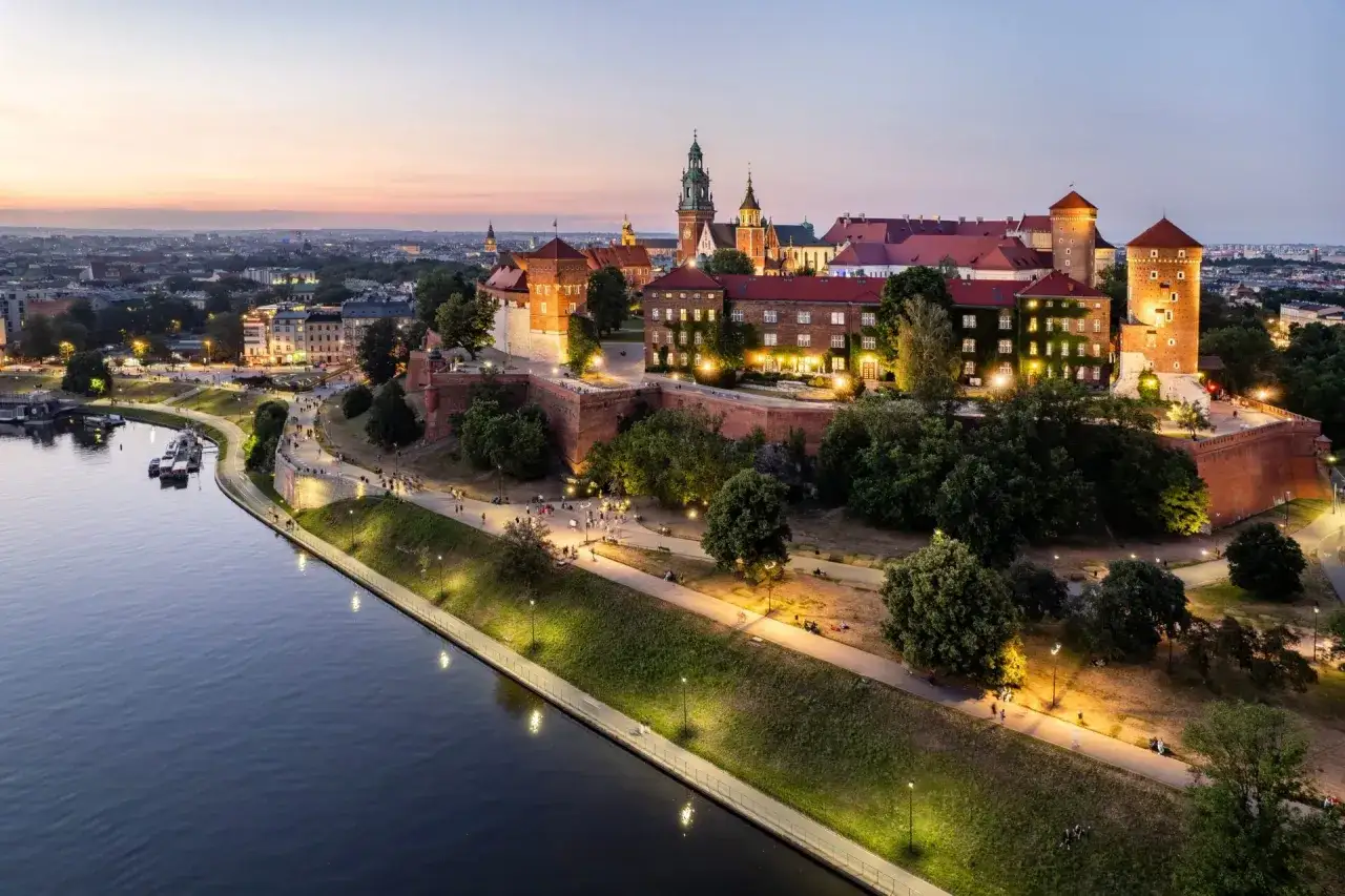 Wieczorne krak&oacute;w zwiedzanie. Wawel lśni w blasku świateł, a Wisła odbija niebo. Spacerowicze cieszą się urokami miasta.