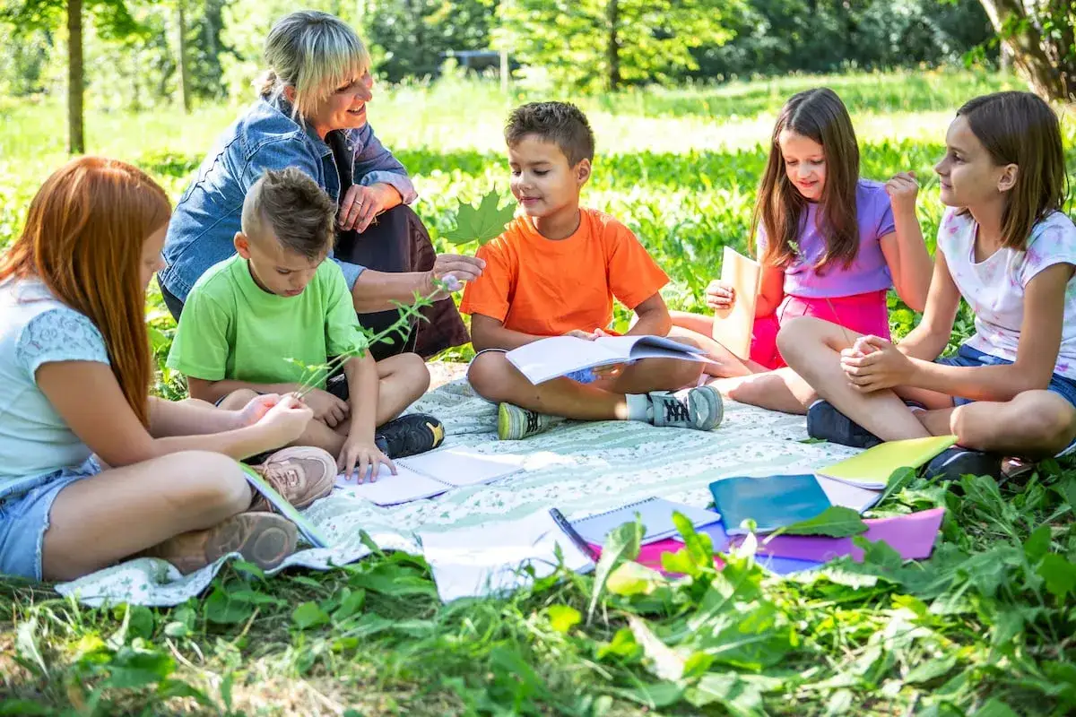 Maestra guía a niños en juegos de conocimiento del medio en un día soleado. Aprenden sobre la naturaleza mientras están sentados en el césped con cuadernos.