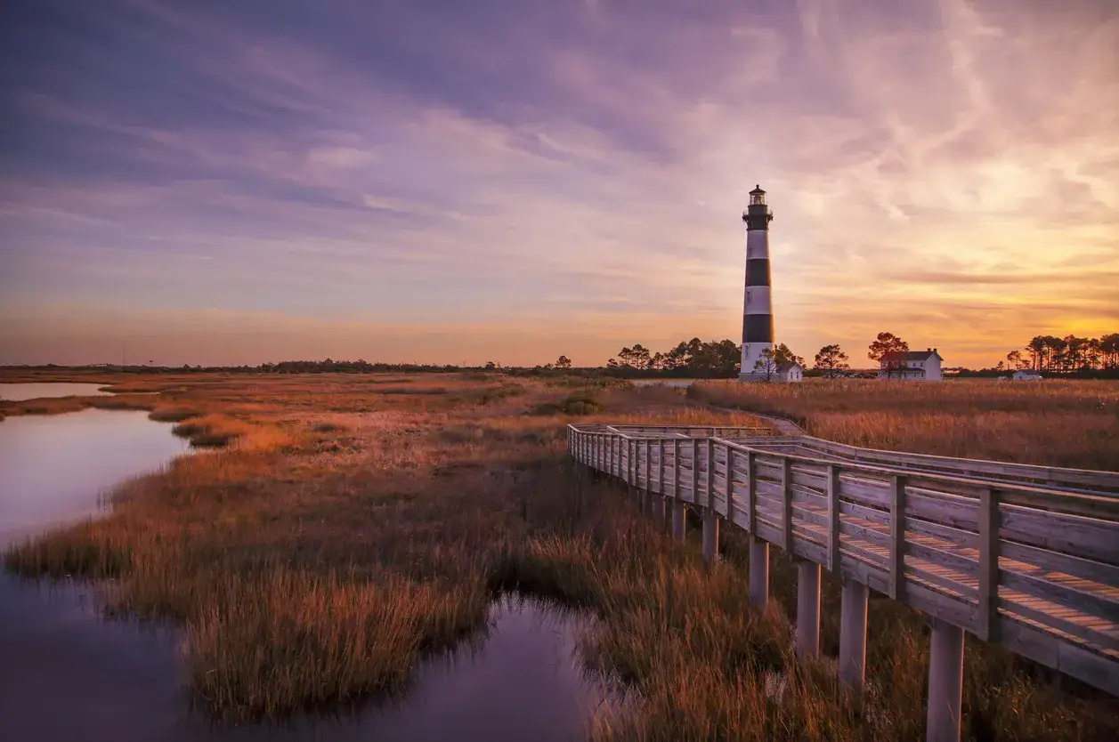 Atardecer en este hermoso outer banks lugar. Un faro icónico se alza sobre marismas doradas, con un camino de madera que invita a explorar.