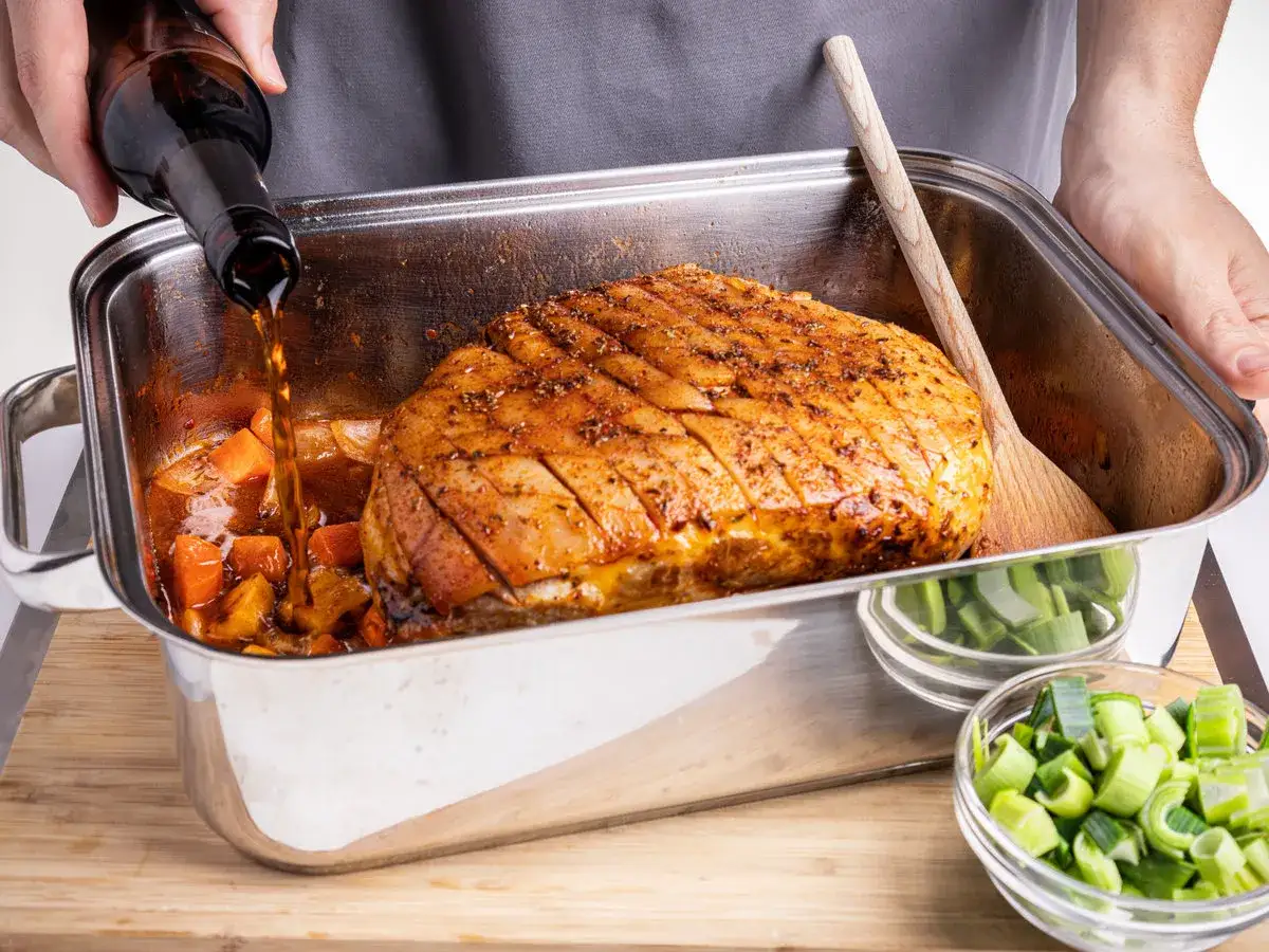 Ein Braten wird in einer Metallform mit Gemüse übergossen. Die richtige schweinebraten temperatur umluft ist entscheidend für ein knuspriges Ergebnis.