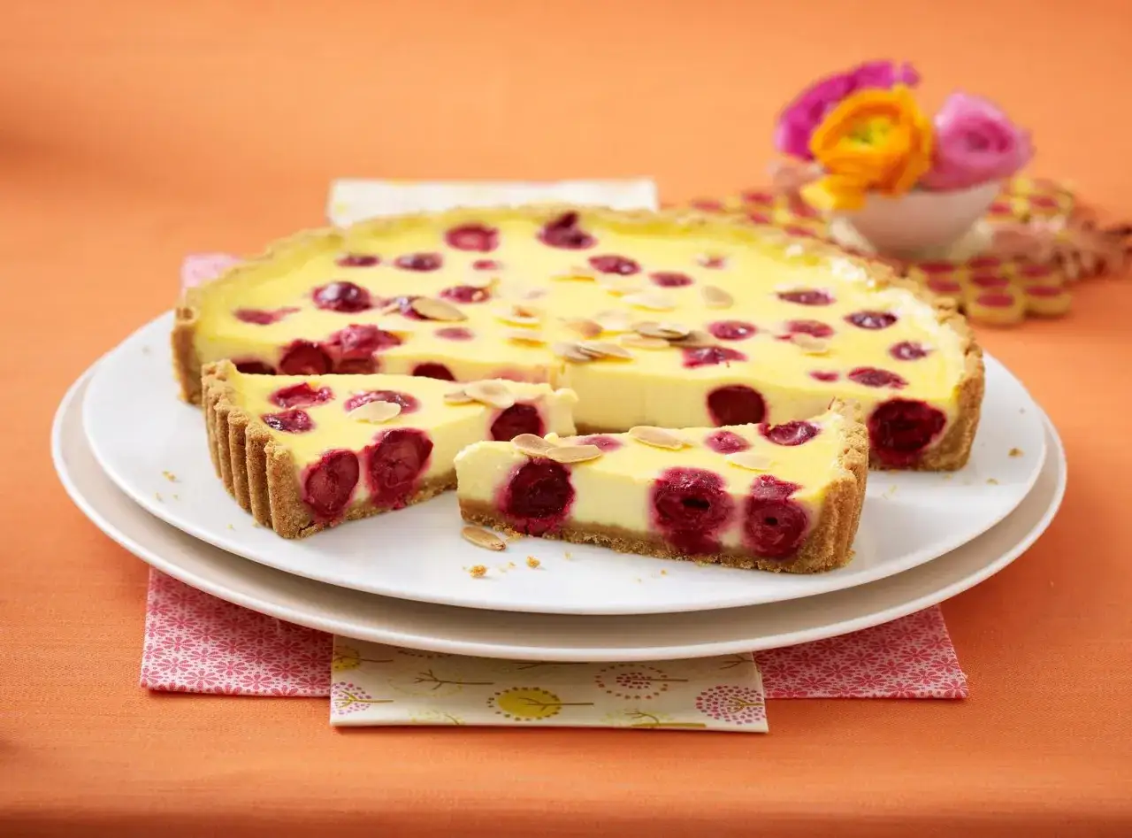 Kuchen mit Keksboden, gefüllt mit Kirschen und Mandelsplittern, auf einem Teller mit bunten Blumen im Hintergrund.