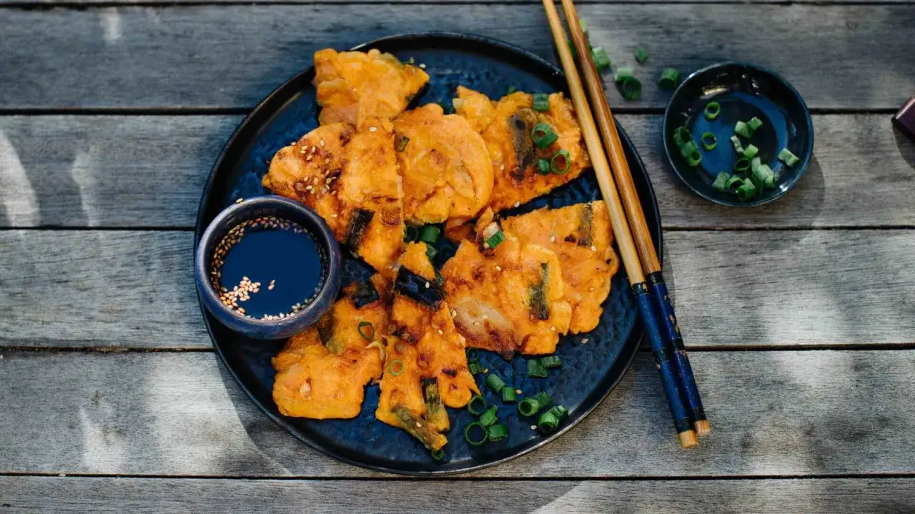 Knusprige Kimchi-Pfannkuchen, garniert mit Fr&uuml;hlingszwiebeln und Sesam, serviert mit Dip-Sauce und St&auml;bchen auf einem Holztisch.