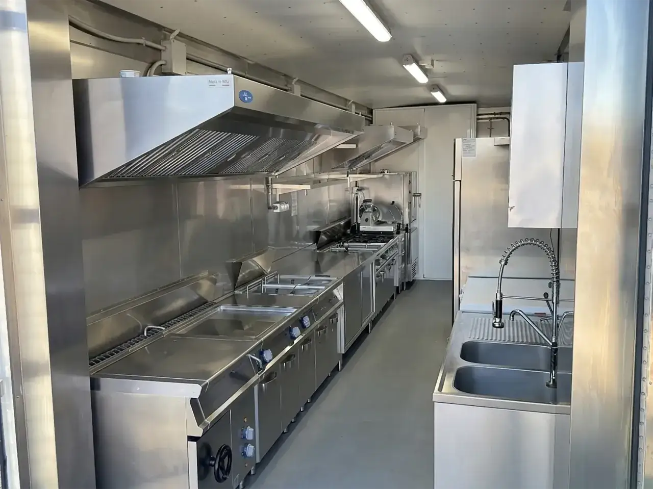 Interno di un food truck con attrezzature professionali in acciaio inox: friggitrici, piastre, cappa aspirante e lavandino doppio.