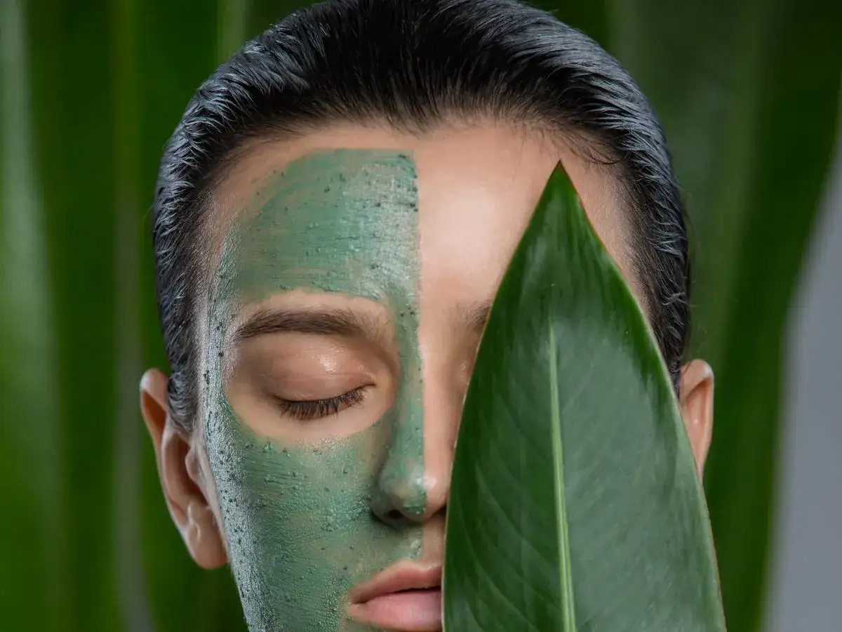 Volto di donna con maschera all'argilla verde, che mostra a cosa serve per una pelle purificata e luminosa, con una foglia verde a coprire parte del viso.