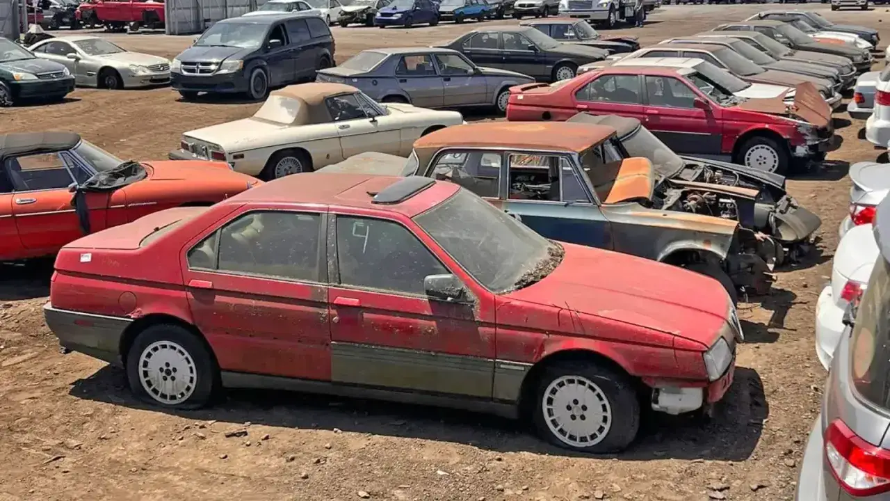 En r&ouml;d Alfa Romeo, ett klassiskt bilm&auml;rke, st&aring;r p&aring; en skrotupplag bland andra bilar.