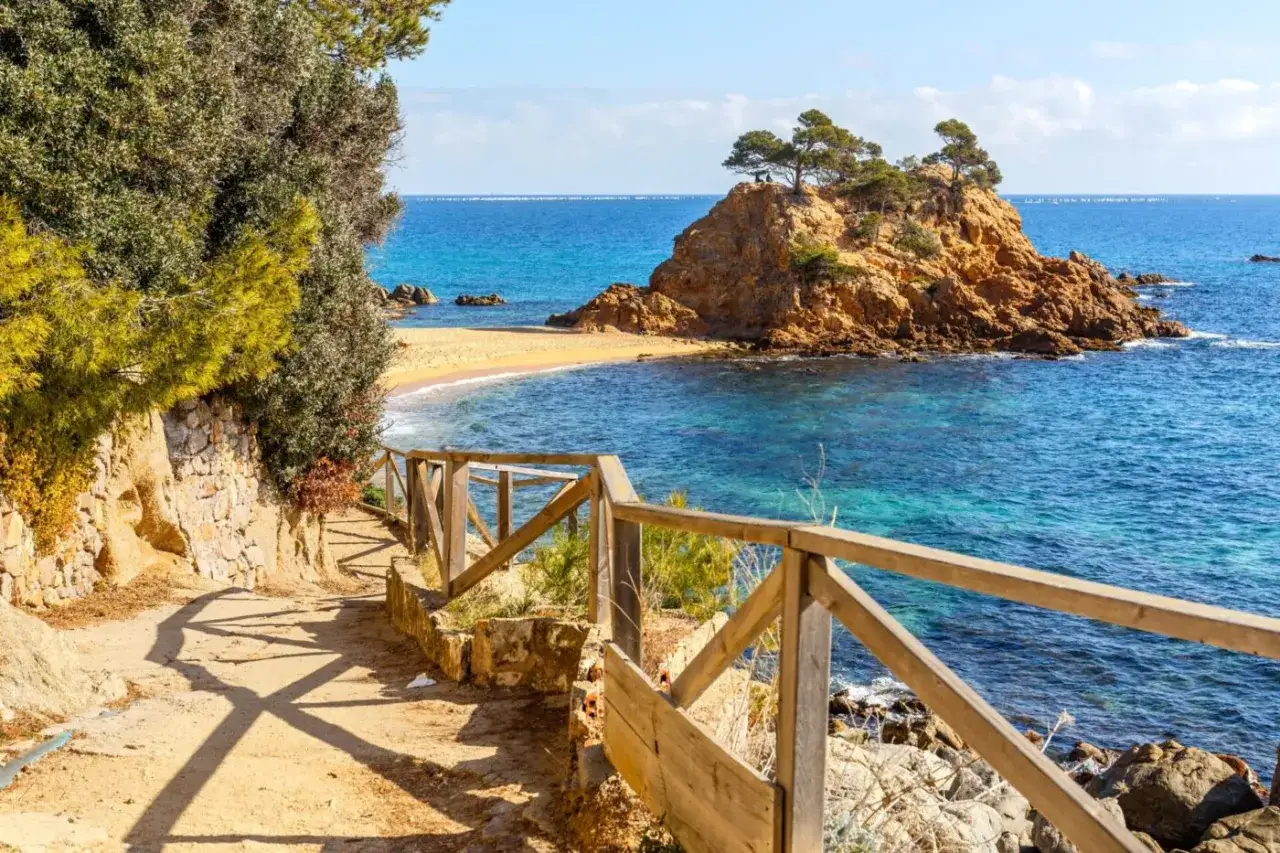 Piękna, słoneczna **costa brava pogoda listopad**. Ścieżka z drewnianym płotkiem prowadzi do zatoczki z turkusową wodą i skalistą wysepką.