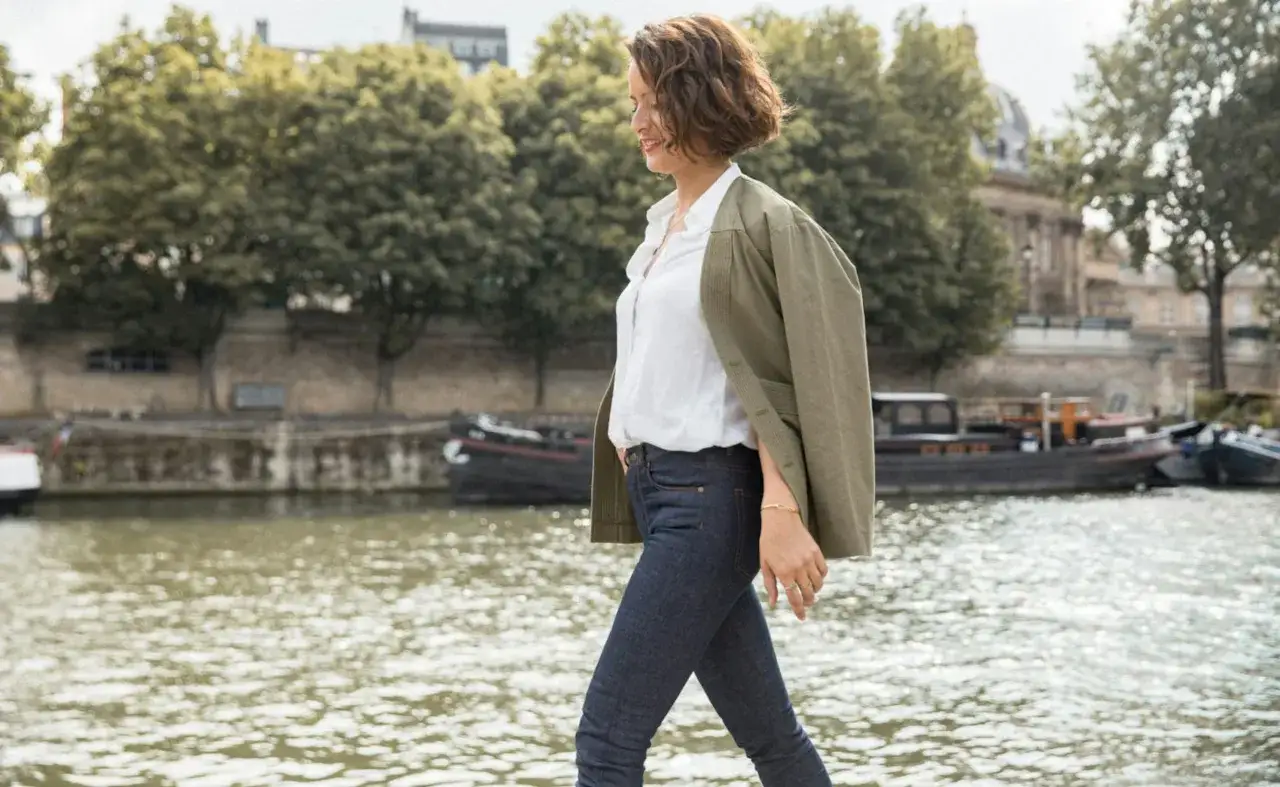 Une femme en jean et chemise blanche, veste kaki, incarne la garde robe idéale pour une balade parisienne au bord de l'eau.