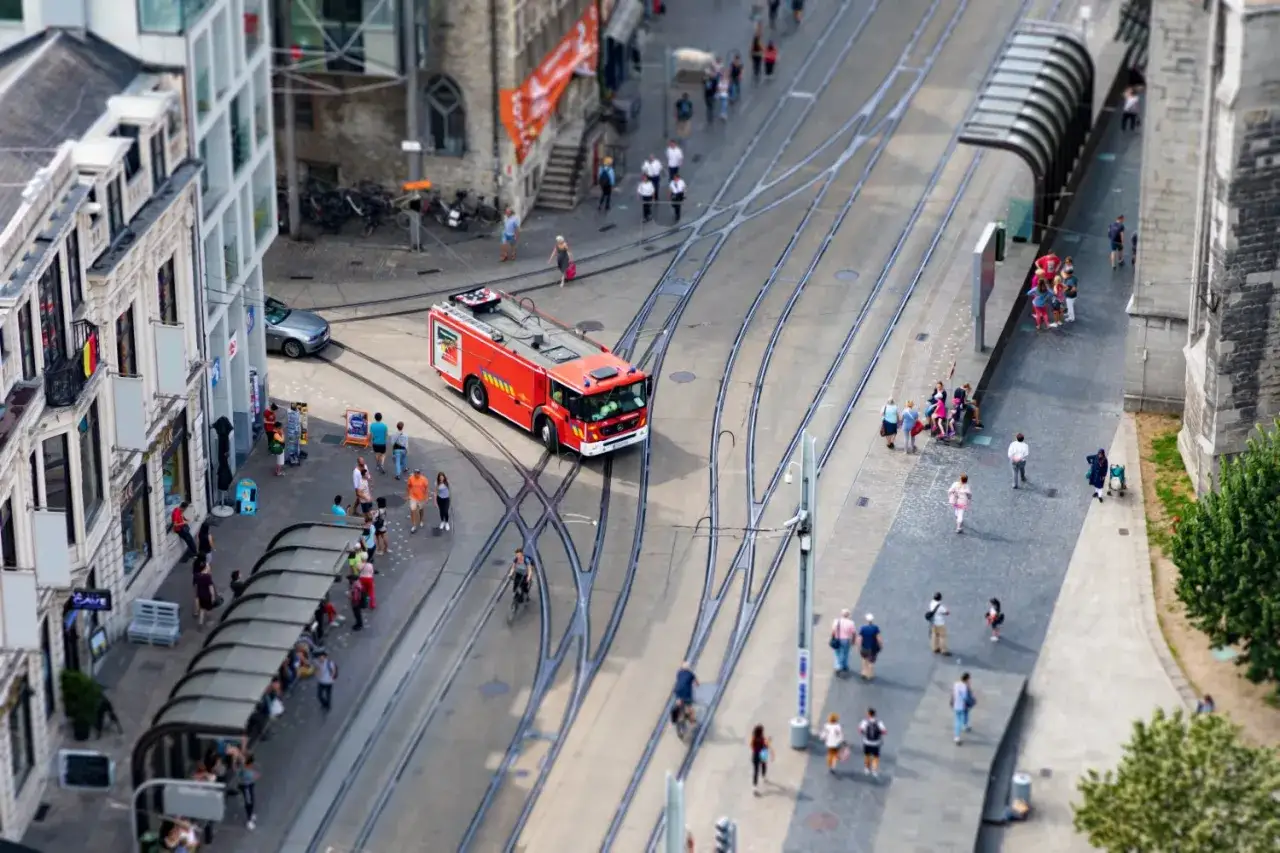 A red fire truck drives through a city street, with people and buildings blurred in the background, creating a miniature effect typical of tilt shift photography.
