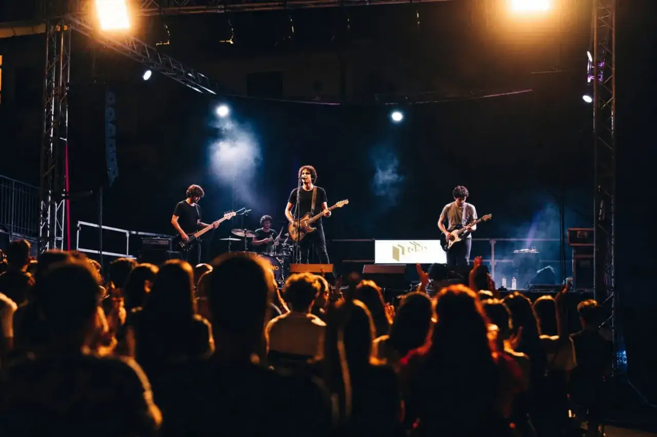 Una tirock band suona sul palco illuminato, con il pubblico che applaude.