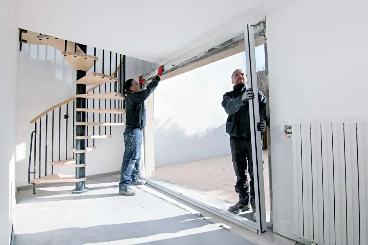 Deux ouvriers effectuent la pose d'une porte fenêtre coulissante. Un escalier hélicoïdal en bois et métal est visible à gauche.
