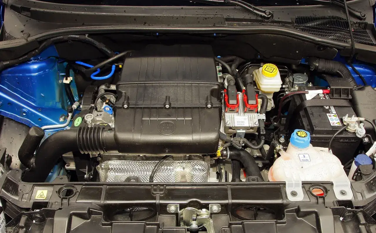 Silnik fiat grande punto 1.4. Widoczny jest czarny plastikowy kolektor dolotowy z logo Fiat, cewki zapłonowe, zbiornik płynu chłodniczego i akumulator.