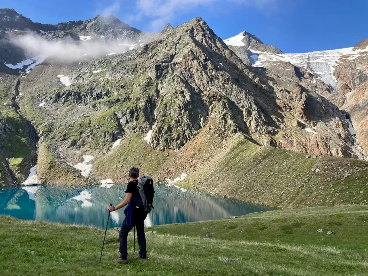 Un randonneur admire un lac bleu vif lors d'un trek en Autriche, avec des montagnes imposantes et des glaciers en arrière-plan.