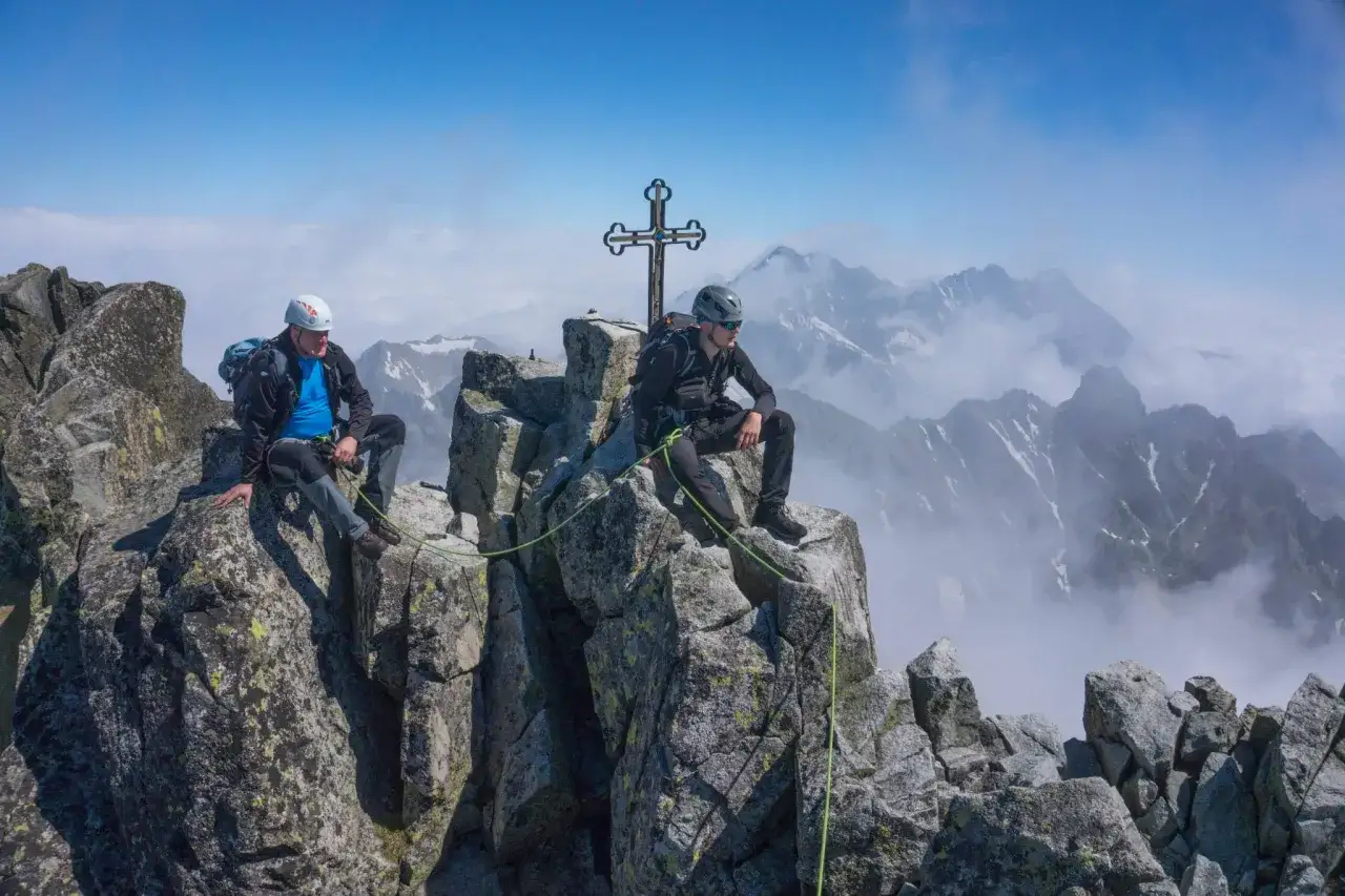 Dw&oacute;ch alpinist&oacute;w odpoczywa na szczycie. W tle rozciąga się majestatyczny widok z polski na Tatry Wysokie, z charakterystycznym krzyżem.