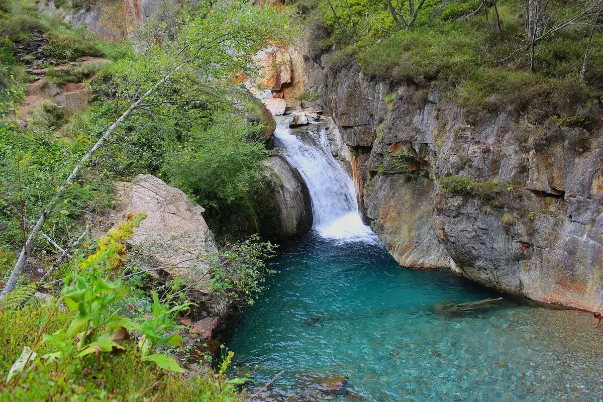La cascade de l'Artigue dévale dans un bassin d'eau turquoise, entourée de roches abruptes et de végétation luxuriante.