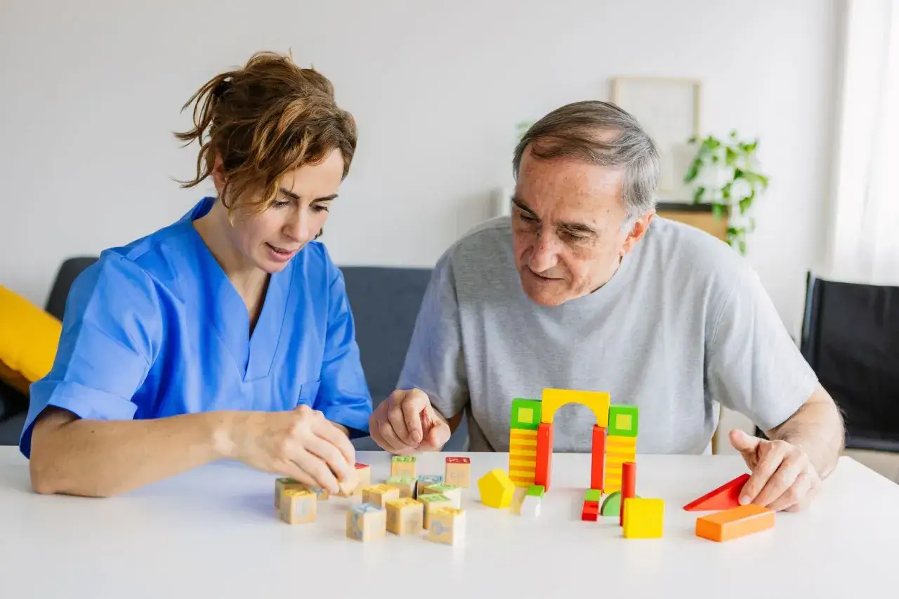 Une aide-soignante en blouse bleue joue avec des cubes en bois avec un homme âgé. Une scène de stimulation cognitive, loin d'un pasa ehpad.