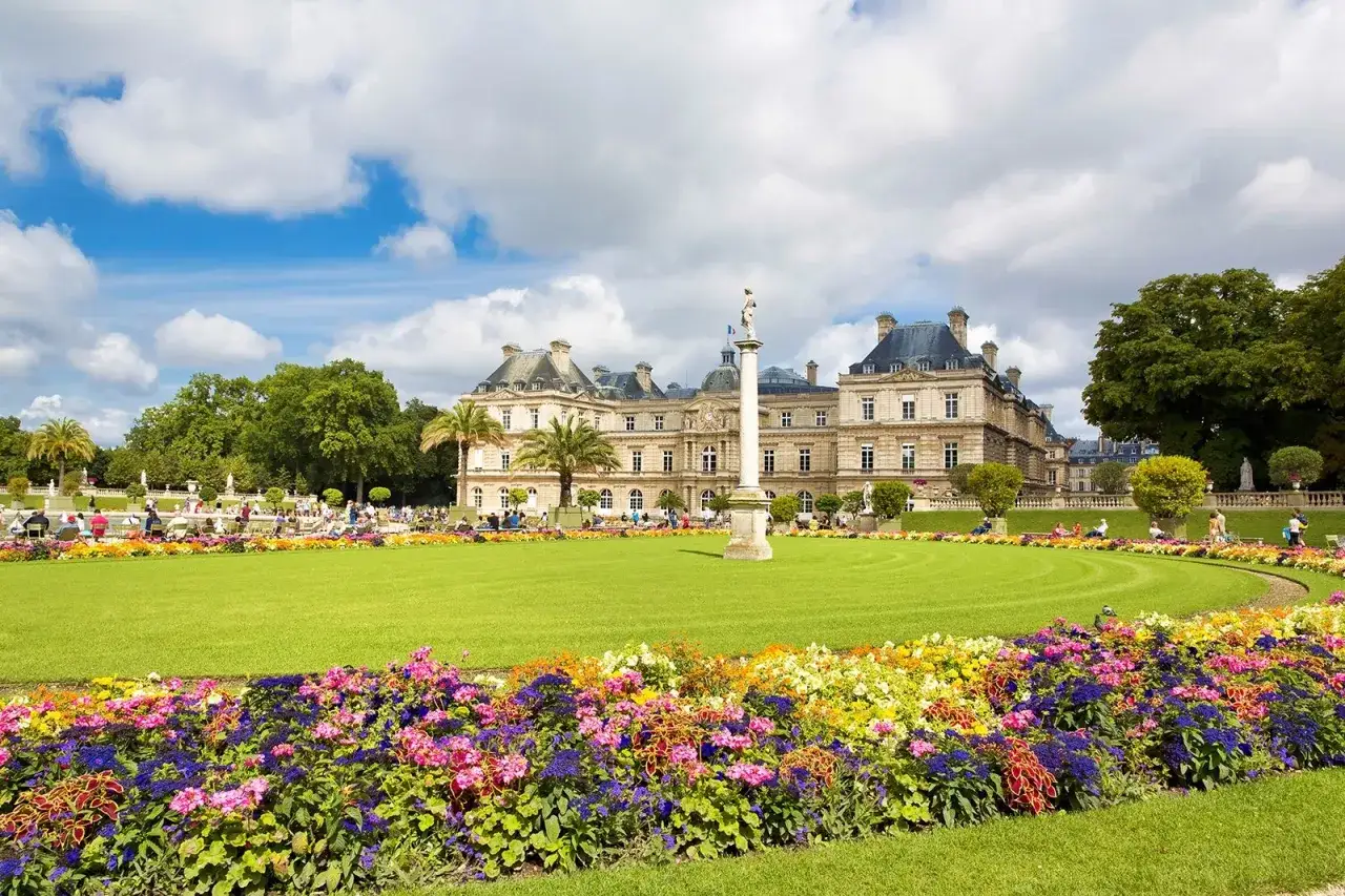 Un d&iacute;a soleado en los jardines de Luxemburgo, con un c&eacute;sped verde impecable, flores vibrantes y el palacio de fondo.