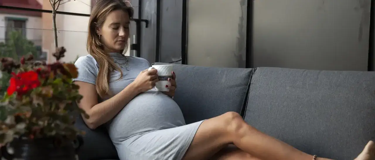 En gravid kvinna i tredje trimestern sitter på en soffa med en kopp i handen.