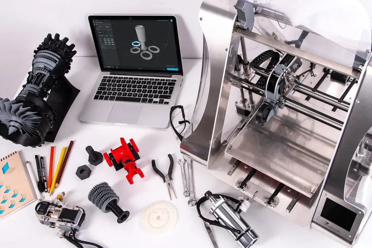 Un operatore CNC progetta e stampa componenti complessi. Sul tavolo, un laptop mostra un modello 3D, accanto a una stampante 3D, parti stampate e attrezzi.