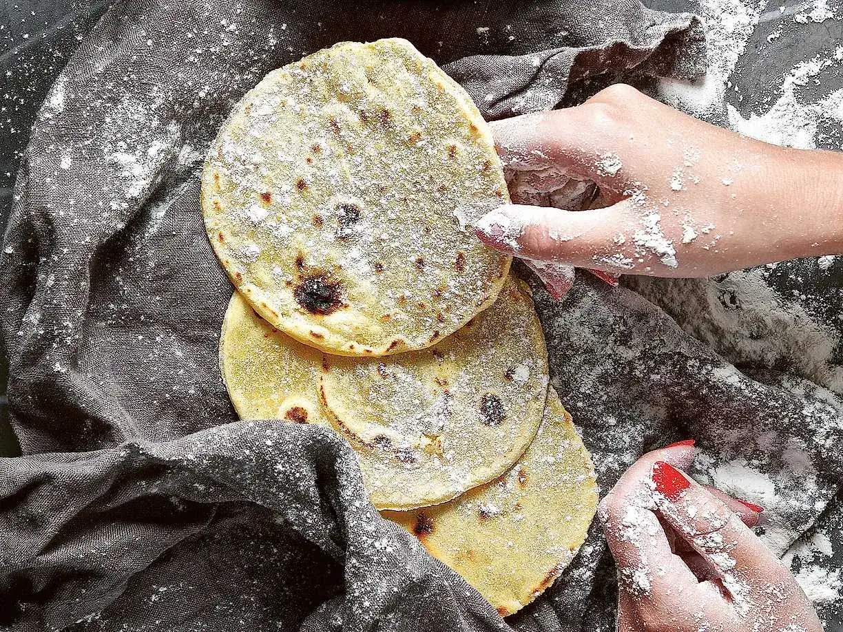 Här är ett enkelt tortilla recept. Flera nybakade tortillas ligger på en grå duk, pudrade med mjöl. Händer med mjöl på fingrarna sträcker sig efter dem.