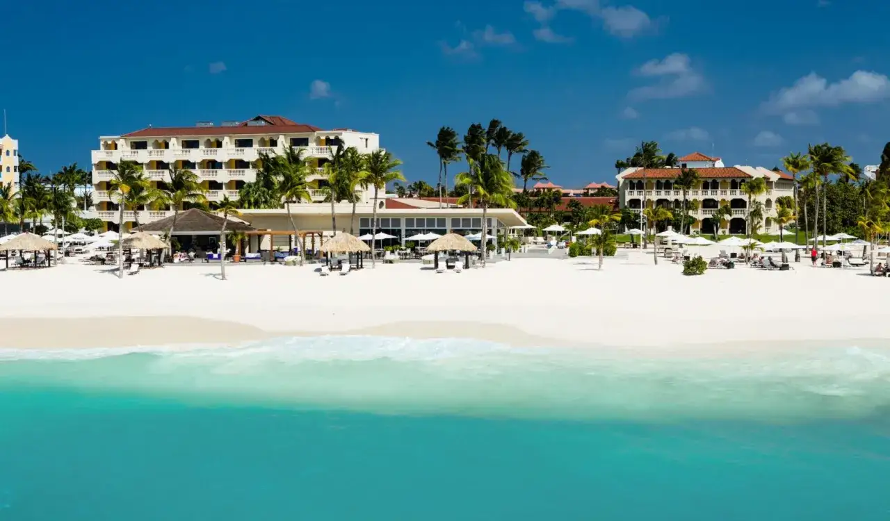 A beautiful white sand beach with turquoise water and palm trees. Hotels line the shore, suggesting a welcoming atmosphere for all travelers, including those asking if Aruba gay friendly.