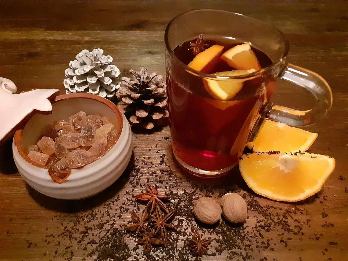 Tee mit Rum, norddeutsch: Ein Glas dunkler Tee mit Orangenscheiben, daneben brauner Kandiszucker, Sternanis, Muskatnüsse und Tannenzapfen auf Holz.