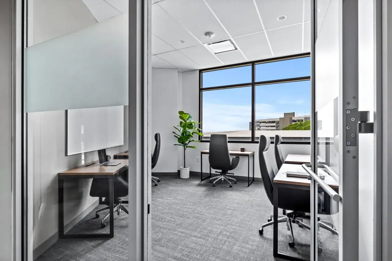 Un bureau flex office moderne avec des postes de travail individuels, une grande fenêtre offrant une vue dégagée et une plante verte.
