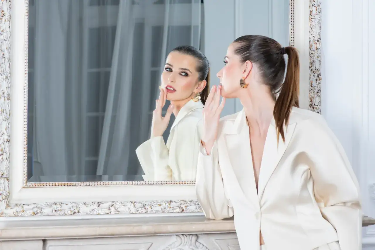 Elegancka kobieta w kremowym żakiecie przegląda się w lustrze. Idealny makijaż i fryzura, doskonałe na szkolenia beauty.