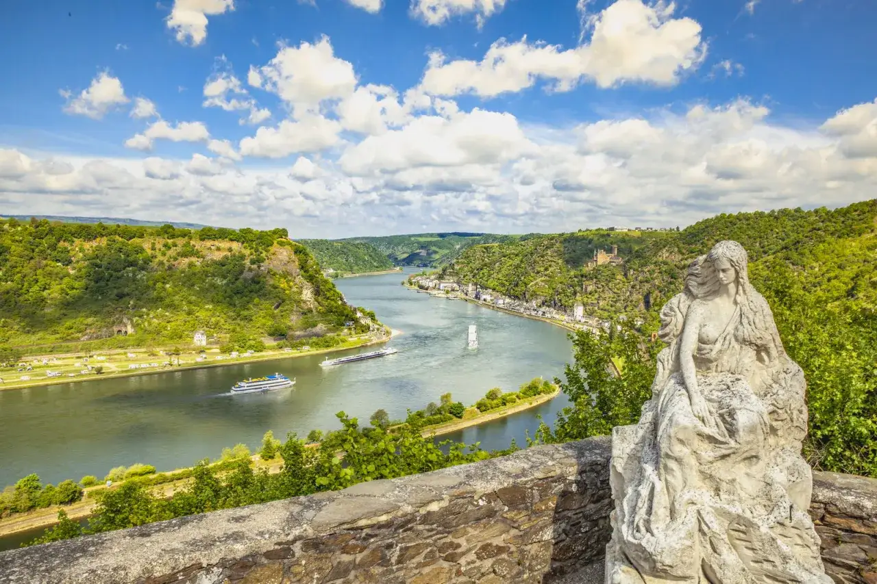 Estatua de mujer junto al río Rin, ruta EuroVelo 15. Barcos navegan por el valle verde.