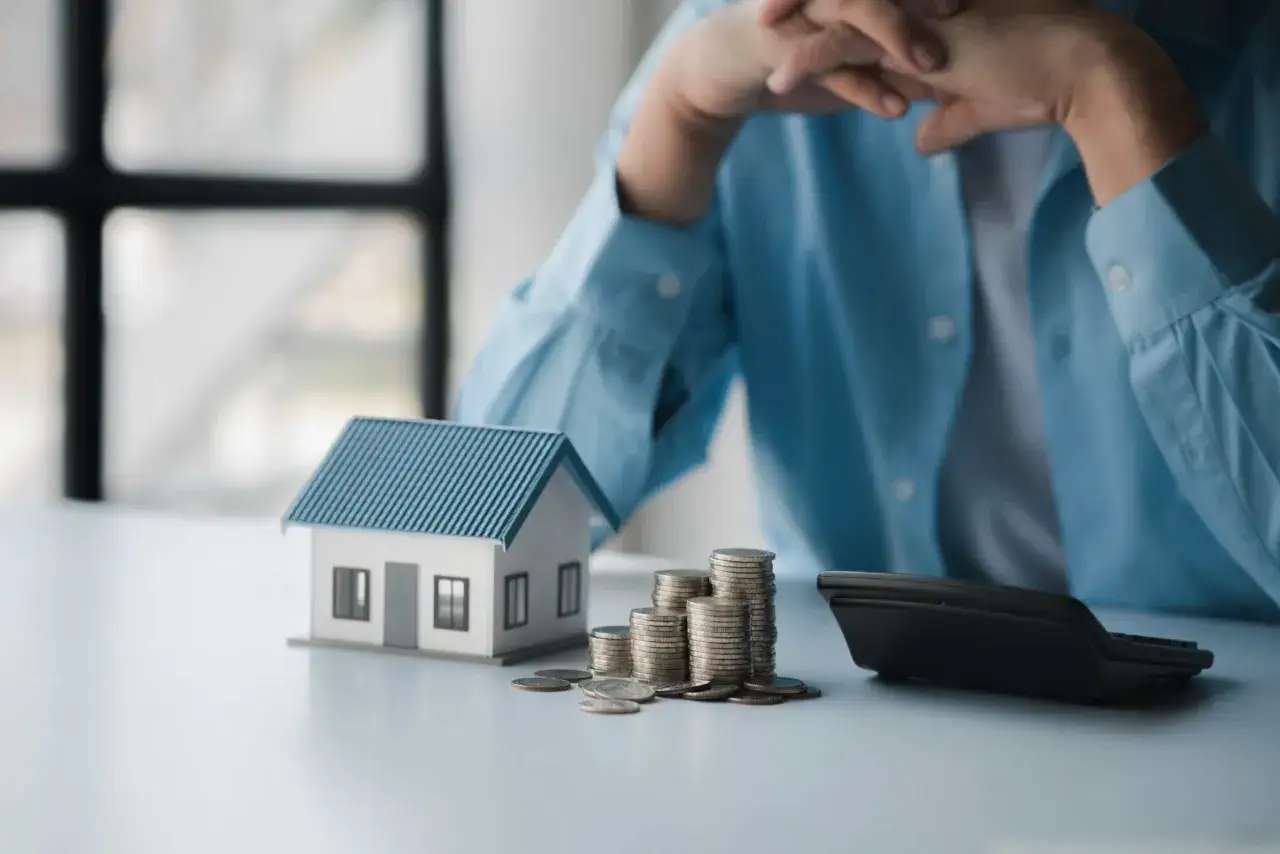 En person funderar över sitt sparande per kvadratmeter i en bostadsrättsförening. En modell av ett hus, mynt och en kalkylator ligger på bordet.
