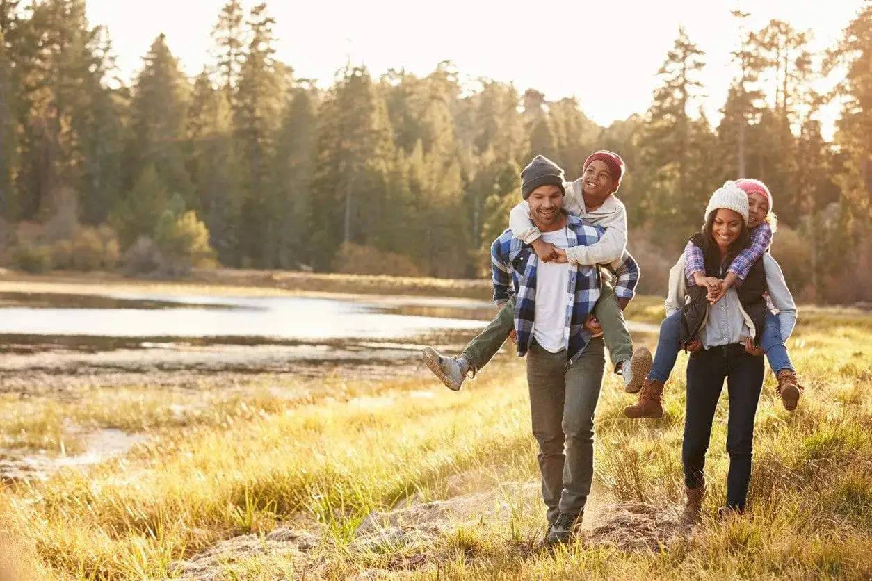 Un couple porte ses enfants sur son dos, profitant d'une micro aventure en famille dans un paysage naturel baigné de soleil.