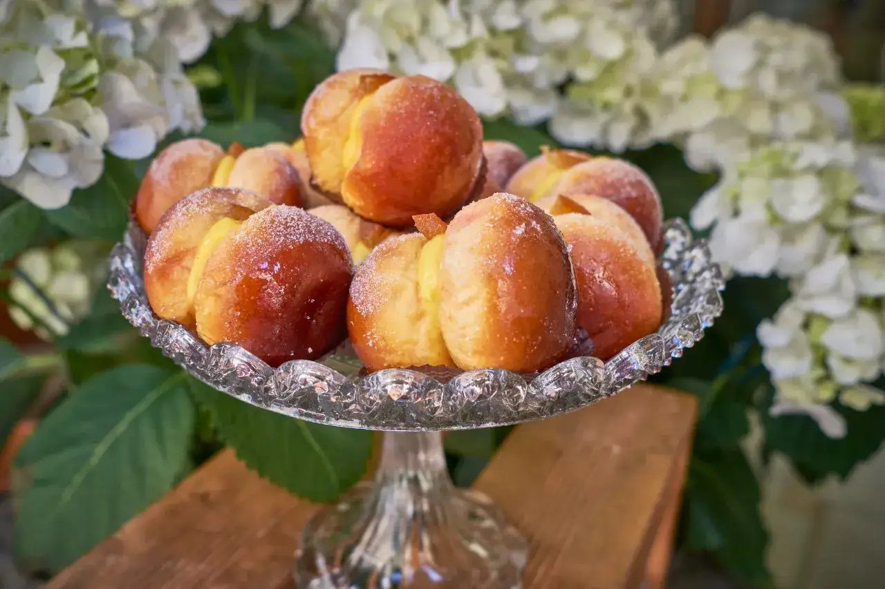 Dolci pesche di prato, ricoperte di zucchero, su un'alzata di cristallo, con fiori bianchi sullo sfondo.