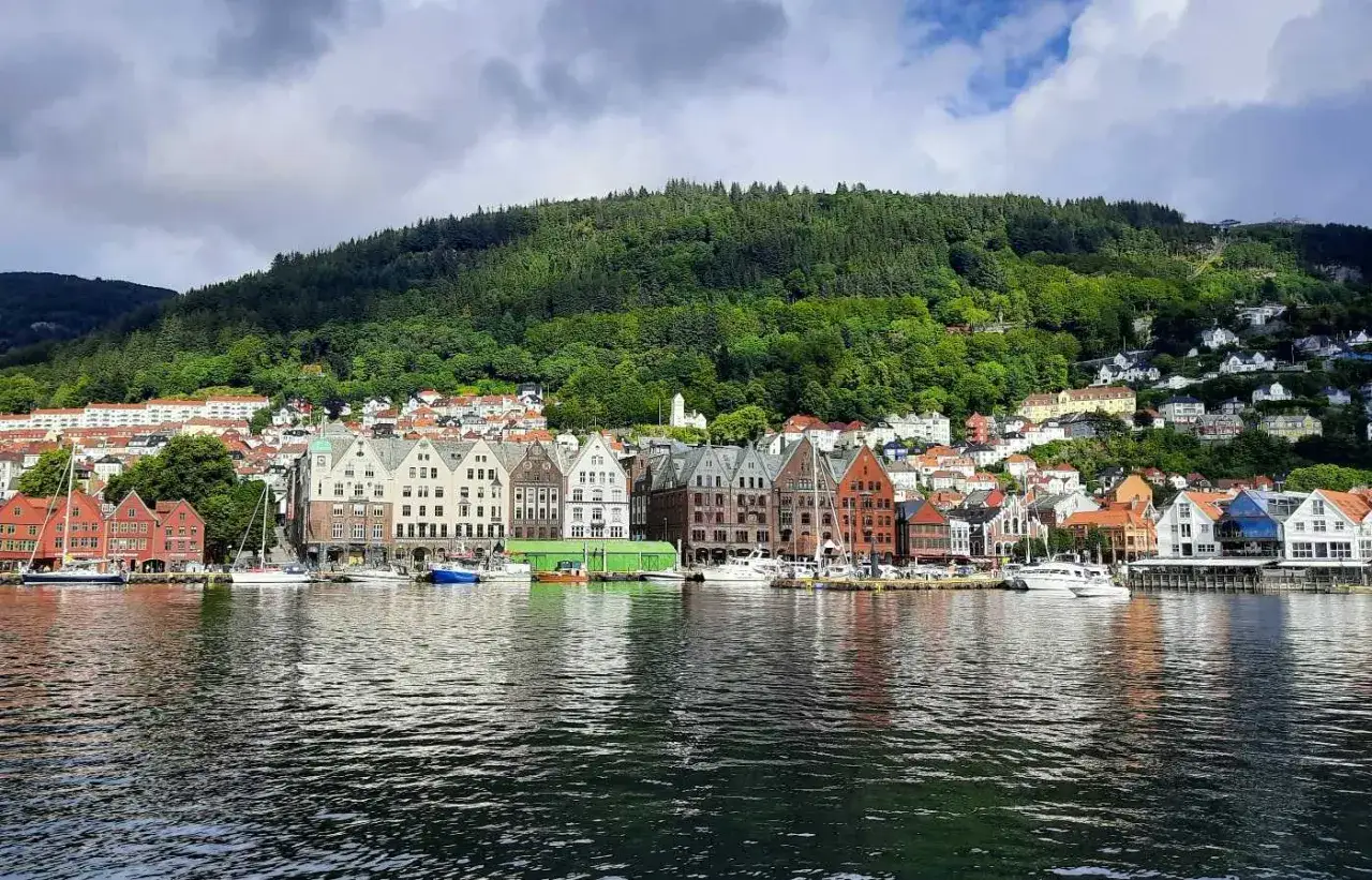 Kolorowe kamienice nad wodą, w tle zielone wzg&oacute;rza z domami. Klasyczny widok na **Bergen, Norwegia**.