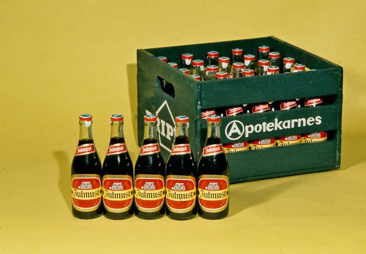 Fem flaskor julmust står framför en grön trälåda fylld med fler flaskor. En del av lådans historia.