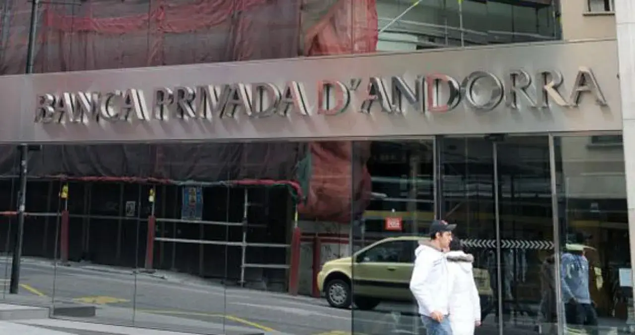 Fachada de Banca Privada d'Andorra, un edificio con letrero metálico y cristales. Personas caminan por la calle, reflejándose en los cristales. BPA finanzas.