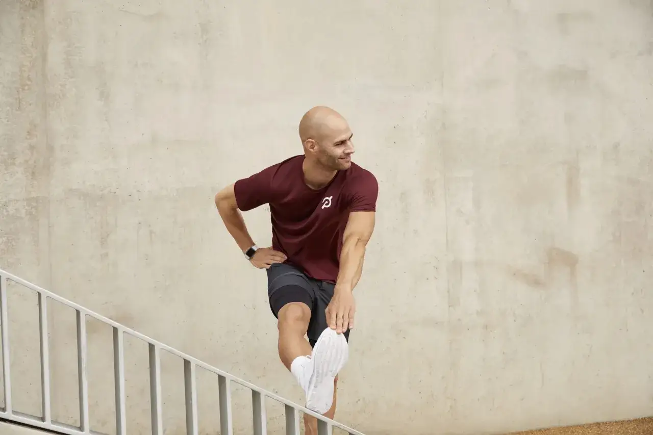 Man in maroon shirt and shorts performs an outer hamstring stretch on stairs, smiling.