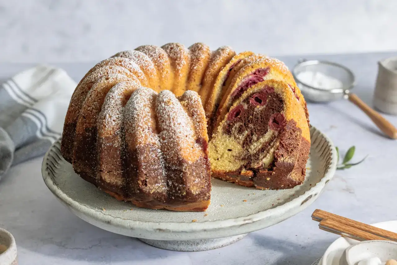 Ein saftiger Marmorkuchen mit Kirschen, bestäubt mit Puderzucker, auf einem grauen Tortenständer.